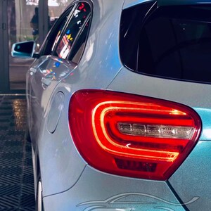 May include: A light blue metallic car with a glossy finish, featuring a prominent red LED taillight assembly with a curved light bar design. The vehicle body displays a smooth, reflective surface, and a stylized logo reading EZ Auto Wrap.