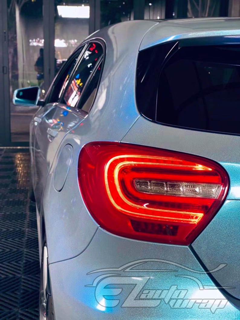 May include: A light blue metallic car with a glossy finish, featuring a prominent red LED taillight assembly with a curved light bar design. The vehicle body displays a smooth, reflective surface, and a stylized logo reading EZ Auto Wrap.