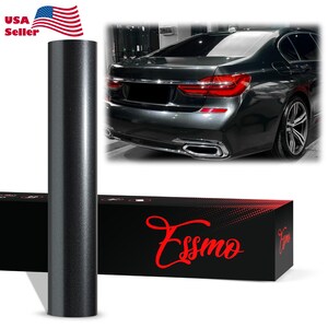 May include: A roll of dark charcoal gray vinyl wrap film standing vertically next to a black rectangular box with the red text Essmo printed on the front. The background features a dark gray sedan showing the rear taillights and exhaust pipes.