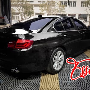 May include: A black sedan with a visible carbon fiber weave pattern on the exterior body panels. The vehicle features silver multi-spoke wheels, red taillights, and dual exhaust pipes. The word Essmo is written in red script in the foreground.