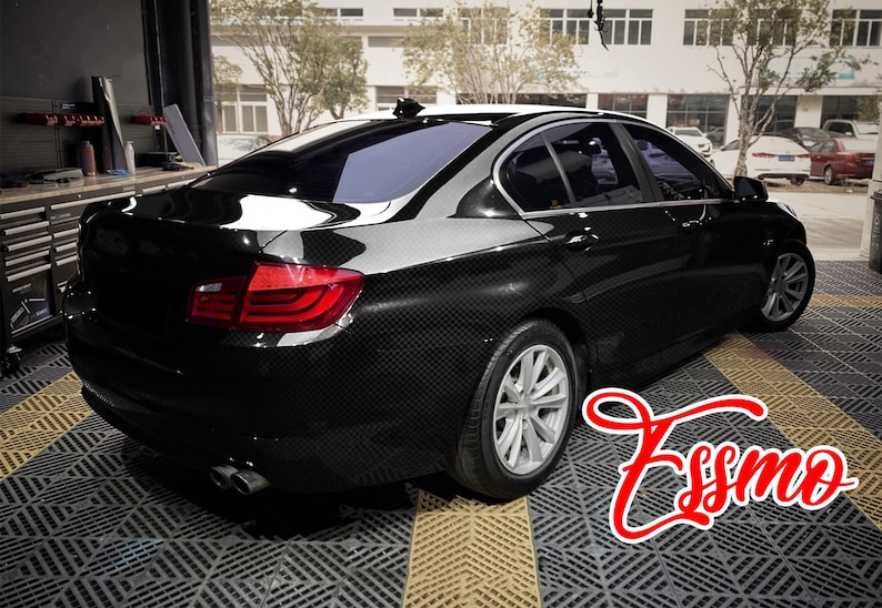 May include: A black sedan with a visible carbon fiber weave pattern on the exterior body panels. The vehicle features silver multi-spoke wheels, red taillights, and dual exhaust pipes. The word Essmo is written in red script in the foreground.