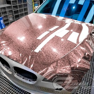 May include: A close-up of a car's hood wrapped in a rose gold metallic vinyl wrap. The wrap has a textured, glittery finish. The hood is partially covered with white tape.