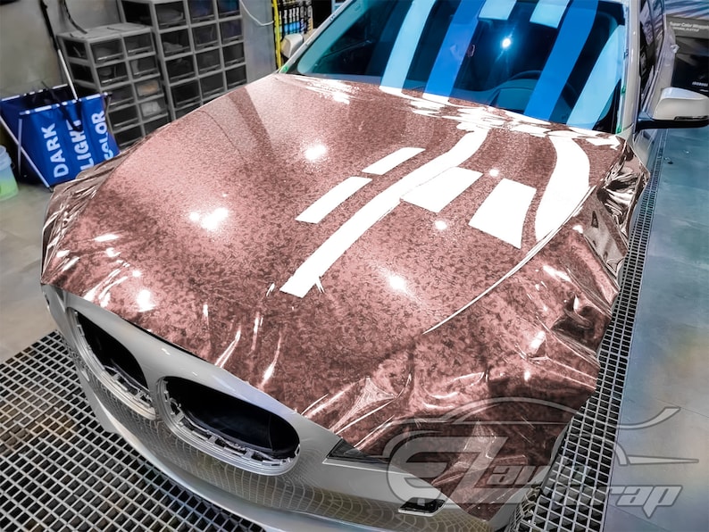 May include: A close-up of a car's hood wrapped in a rose gold metallic vinyl wrap. The wrap has a textured, glittery finish. The hood is partially covered with white tape.