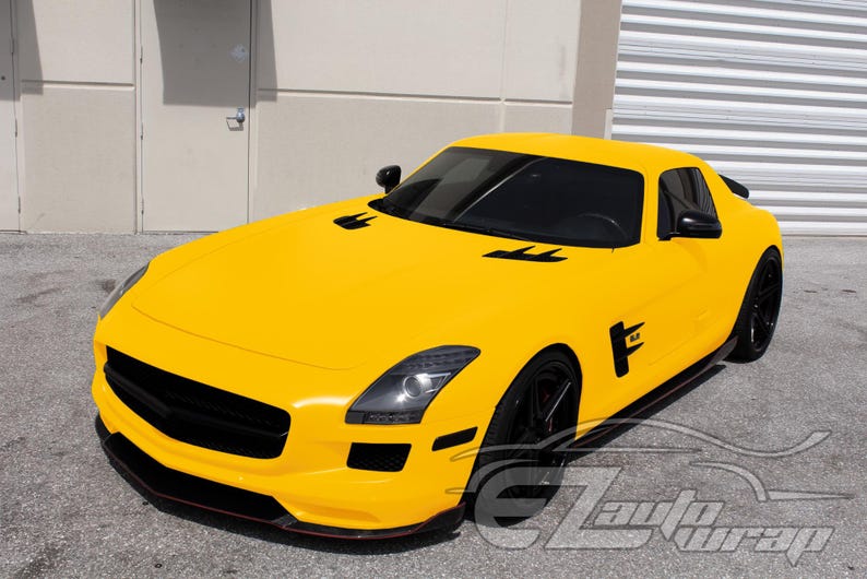 May include: A bright yellow sports car featuring a sleek aerodynamic body, black front grille, black side mirrors, and black multi-spoke wheels. The vehicle has tinted windows and black air vents on the hood. The text EZAutoWrap is visible.