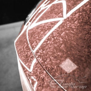 May include: Close-up of a rose gold car wrap with a carbon fiber pattern. The wrap has a glossy finish and is applied to the hood of the car.