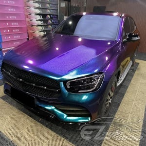 May include: A luxury SUV featuring a color-shifting iridescent wrap that transitions between deep purple, vibrant blue, and teal tones. The vehicle has a black mesh front grille, sleek LED headlights, and a glossy finish that reflects light.