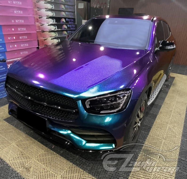 May include: A luxury SUV featuring a color-shifting iridescent wrap that transitions between deep purple, vibrant blue, and teal tones. The vehicle has a black mesh front grille, sleek LED headlights, and a glossy finish that reflects light.