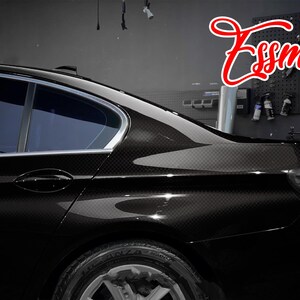 May include: A black sedan featuring a carbon fiber wrap finish on the rear quarter panel and door. The vehicle has tinted windows and a silver trim along the roofline. The red script text Essmo is visible in the upper right corner of the image.