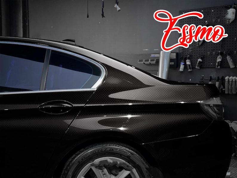 May include: A black sedan featuring a carbon fiber wrap finish on the rear quarter panel and door. The vehicle has tinted windows and a silver trim along the roofline. The red script text Essmo is visible in the upper right corner of the image.