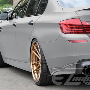 May include: A matte gray sedan featuring custom metallic bronze multi-spoke wheels, a rear spoiler, and quad exhaust tips. The vehicle displays a sleek, low-profile design with tinted windows and a watermark logo reading EZAutoWrap in the corner.