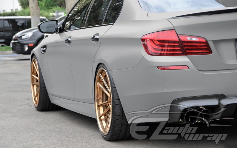 May include: A matte gray sedan featuring custom metallic bronze multi-spoke wheels, a rear spoiler, and quad exhaust tips. The vehicle displays a sleek, low-profile design with tinted windows and a watermark logo reading EZAutoWrap in the corner.