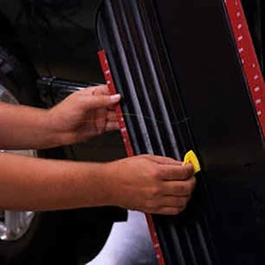 May include: A person's hands are shown applying a red adhesive tape to a black surface. The tape is being smoothed down with a yellow tool.