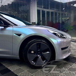 May include: A Tesla Model 3 sedan featuring a custom iridescent pearlescent wrap that shifts between silver, blue, and purple hues. The vehicle includes dark grey aerodynamic wheel covers, black side mirror caps, and a sleek, modern front bumper design.