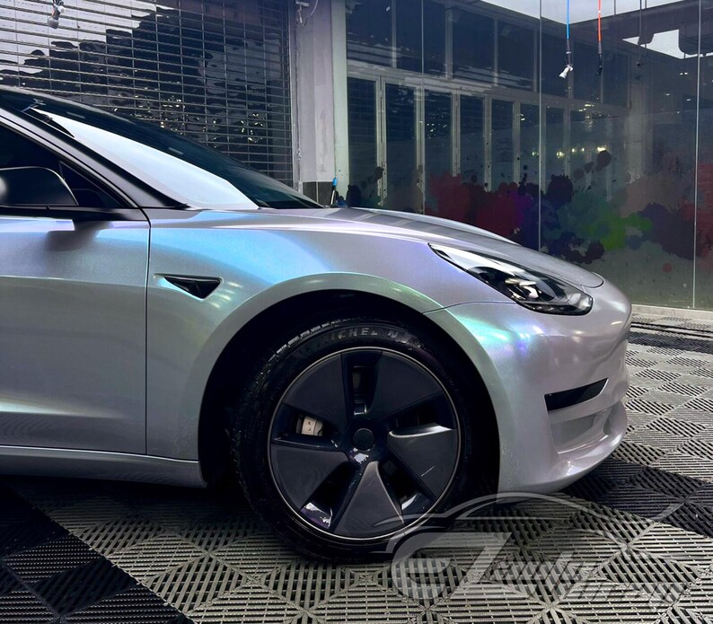 May include: A Tesla Model 3 sedan featuring a custom iridescent pearlescent wrap that shifts between silver, blue, and purple hues. The vehicle includes dark grey aerodynamic wheel covers, black side mirror caps, and a sleek, modern front bumper design.