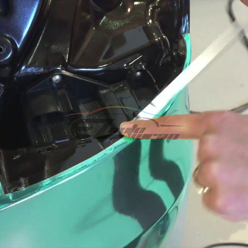 May include: A close-up of a car being wrapped in a teal green vinyl wrap. A person's hand is holding a squeegee to smooth out the vinyl.