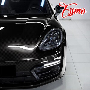 May include: A black car with a visible carbon fiber weave pattern on the hood, featuring a sleek headlight assembly, a lower air intake with a horizontal LED light strip, and the red cursive text Essmo positioned in the upper right corner.