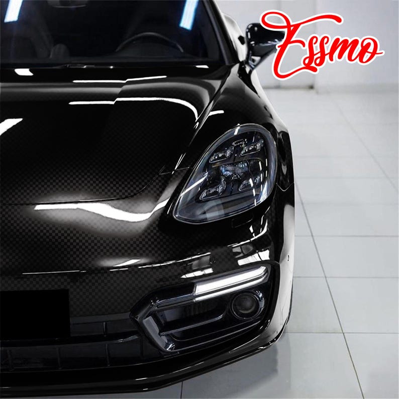 May include: A black car with a visible carbon fiber weave pattern on the hood, featuring a sleek headlight assembly, a lower air intake with a horizontal LED light strip, and the red cursive text Essmo positioned in the upper right corner.