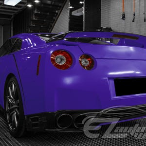 May include: A vibrant purple sports car viewed from the rear three-quarter angle, featuring a prominent rear spoiler, circular taillights, and a quad-exhaust system. The vehicle has a sleek body design with dark alloy wheels and a blacked-out license plate.