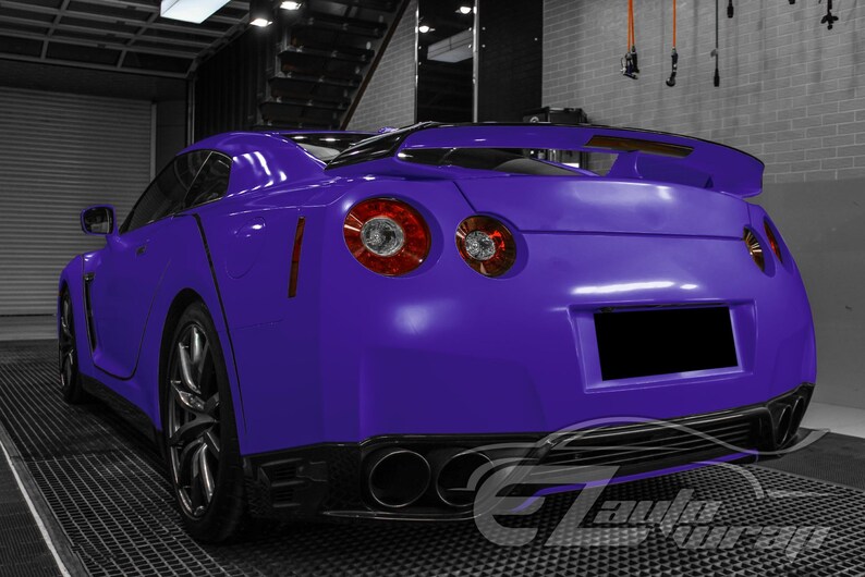 May include: A vibrant purple sports car viewed from the rear three-quarter angle, featuring a prominent rear spoiler, circular taillights, and a quad-exhaust system. The vehicle has a sleek body design with dark alloy wheels and a blacked-out license plate.