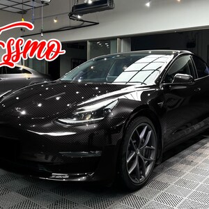 May include: A black sedan with a visible carbon fiber weave pattern on the exterior body panels. The vehicle features dark grey multi-spoke alloy wheels and sleek headlights. The word Essmo is written in a red cursive font in the upper left corner.