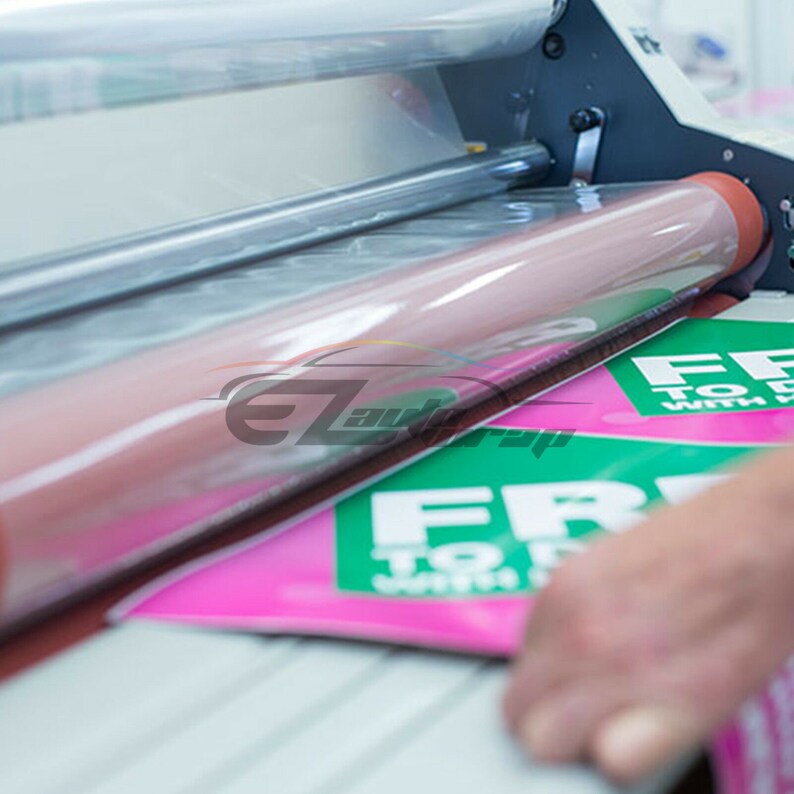 Pu&ograve; includere: Una macchina per plastificare con un foglio di plastica trasparente che viene alimentato attraverso i rulli. La macchina sta plastificando una pila di volantini rosa e verdi che recitano "Gratis per tutti con l'acquisto".
