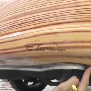 May include: A close-up of a wood grain vinyl wrap being applied to a car. The wrap is a light brown color with darker brown stripes. The wrap is being smoothed out with a squeegee.