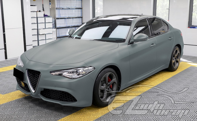 May include: A matte gray Alfa Romeo Giulia sedan featuring a distinctive triangular front grille, sleek headlights, and dark gray alloy wheels with visible red brake calipers. The vehicle has a smooth, non-reflective finish and a black roof panel.