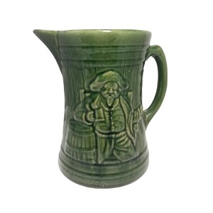 May include: Green ceramic pitcher with a raised design of a man holding a mug and standing next to a barrel.