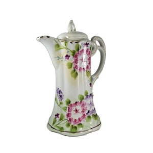 May include: A white porcelain creamer with a floral design. The creamer has a lid and a handle. The flowers are pink, purple, and green.