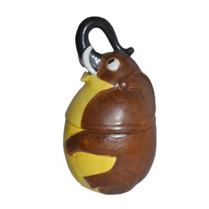 Pode incluir: Um recipiente de cerâmica em forma de elefante, com corpo castanho e barriga amarela. A tromba do elefante é preta e as presas são brancas. O recipiente tem uma tampa e é provavelmente usado para armazenamento ou decoração.