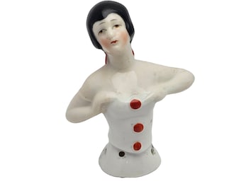 May include: A porcelain doll with black hair and a white dress with red buttons. The doll is missing its arms and is posed with its hands on its chest.