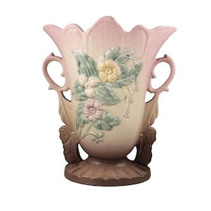 May include: A ceramic vase with a scalloped rim and two side handles. The body features a gradient from light pink at the top to a darker brown base. A raised relief design of green leaves and pastel yellow and pink flowers adorns the front center.