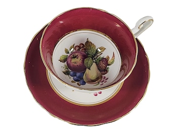 May include: A vintage teacup and saucer set. The cup and saucer are a deep burgundy color with gold trim. The center of the cup and saucer feature a colorful fruit design, including grapes, apples, and pears. A classic tea set.