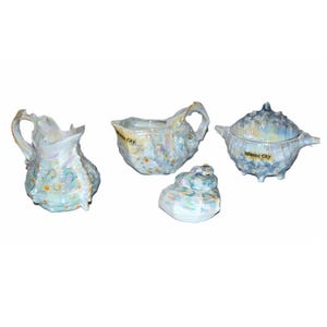 May include: A set of four iridescent ceramic seashell-shaped serving dishes. The set includes a creamer, a sugar bowl with a lid, and two other dishes. The text "Atlantic City" is printed on two of the pieces. The dishes have a pearlescent finish.