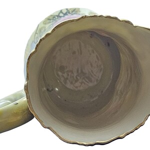 Może przedstawiać: Wazon ceramiczny w stylu vintage z falistym brzegiem i uchwytem. Wazon ma kremowe wnętrze i jasnozielony wygląd zewnętrzny ze złotym wykończeniem. Na zewnątrz widoczne są kwiatowe wzory.
