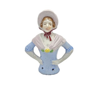 May include: A porcelain doll head with a blue and white dress, a pink hat, and a red bow. The doll has a painted face with brown hair and blue eyes.