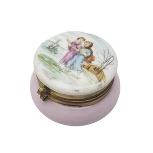 May include: A small, round, pink porcelain trinket box with a hinged lid. The lid features a hand-painted scene of two women walking in a landscape with a tree and a small building. The box has a gold-toned metal hinge.
