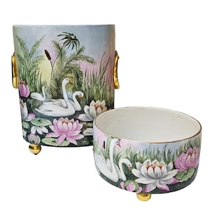 Op de afbeelding: Tweedelige keramische set met een zwaan- en waterlelie-ontwerp. Het hogere stuk heeft gouden handvatten en voetjes. De set toont een handgeschilderde scène met witte zwanen, roze waterlelies en groene bladeren tegen een zachte blauwroze achtergrond.