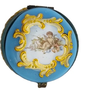 Pode incluir: Pequena caixa redonda para bijuterias com exterior em esmalte azul claro. A tampa apresenta uma cena central pintada de um querubim com pombas, emoldurada por um desenho de filigrana dourada. A caixa tem uma borda de metal escuro.