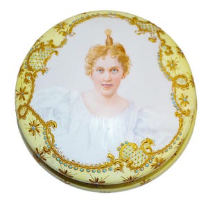 May include: A round, antique porcelain trinket box with a portrait of a woman. The lid is pale yellow with gold filigree and turquoise accents. The portrait features a woman with blonde hair and a white dress.