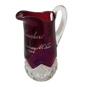 May include: A deep red glass jug with a clear glass handle and a clear, diamond-patterned base. The body features etched white cursive text that reads Vaughn, Martinsburg, W. Va., and 1913, with a curved pouring spout at the top rim.