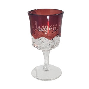 May include: A clear glass goblet with a ruby red base and a cut glass design. The goblet is etched with the words "Oregon City".