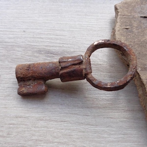 May include: Close-up of a rusty, antique key with a circular handle. The key is brown and shows signs of age and wear. The key is lying on a light gray wooden surface, next to a piece of rough, textured wood.