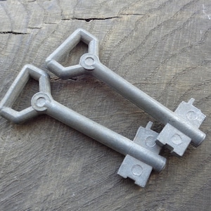 May include: Two silver-colored keys with a geometric handle design and a rectangular key bit. The keys are lying on a weathered, gray wooden surface. The keys have a vintage or antique aesthetic.