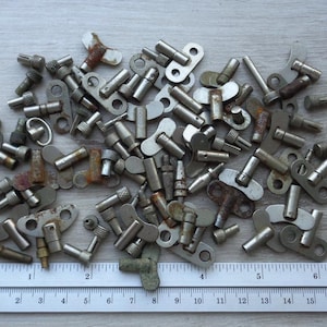 Op de afbeelding: Een verzameling vintage metalen kloksleutels en opwindgereedschap, in verschillende vormen en maten, verspreid over een houten oppervlak. De sleutels zijn zilverkleurig en sommige vertonen roest. Een liniaal met cm-maten staat onderaan.