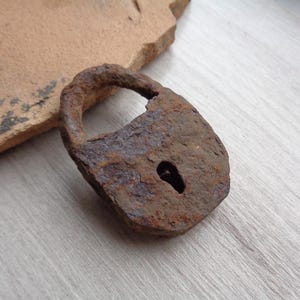 May include: A weathered, antique padlock, showing signs of rust and age. The lock has a keyhole and a curved handle. It is a dark brown colour and rests on a light-coloured wooden surface.