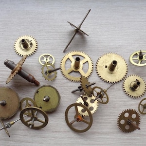 Op de afbeelding: Een verzameling van 13 messing tandwielen en uurwerk onderdelen, waaronder een stervormig tandwiel, een rond tandwiel met een centraal gat en een tandwiel met een lange metalen staaf eraan bevestigd. De tandwielen zijn gerangschikt op een licht houten oppervlak.