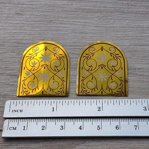 May include: Two arched gold-colored metal plaques featuring a repeating floral and vine pattern in white. A metal ruler is positioned below the plaques, showing each piece is approximately 1.5 inches wide and 1.75 inches tall at the highest point.
