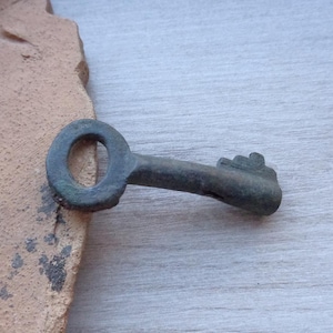 May include: A small, antique-style key with a circular handle and a detailed bit. The key is made of a dark, aged metal, possibly bronze or iron, with a slightly weathered appearance. The key is lying on a light-colored wooden surface.