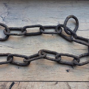 以下が含まれることがあります： 暗く風化した錆びた茶色の仕上げが施された、相互に連結された楕円形のリンクからなる頑丈な金属チェーン。チェーンは緩やかな曲線を描くように配置されており、すべての金属部品に粗い酸化した質感が現れています。